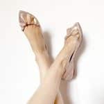 Woman wearing rose gold ballet flats with ruched bow detail – legs crossed against white background